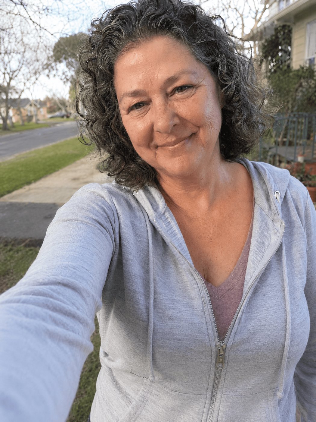 A natural, slightly imperfect selfie-style outdoor portrait of a happy, healthy woman ~54 with a slim face, real skin and a warm genuine smile, wearing a casual zip-up jacket, with a suburban street or yard slightly blurred behind her. Looks like a real photo a real woman would post.